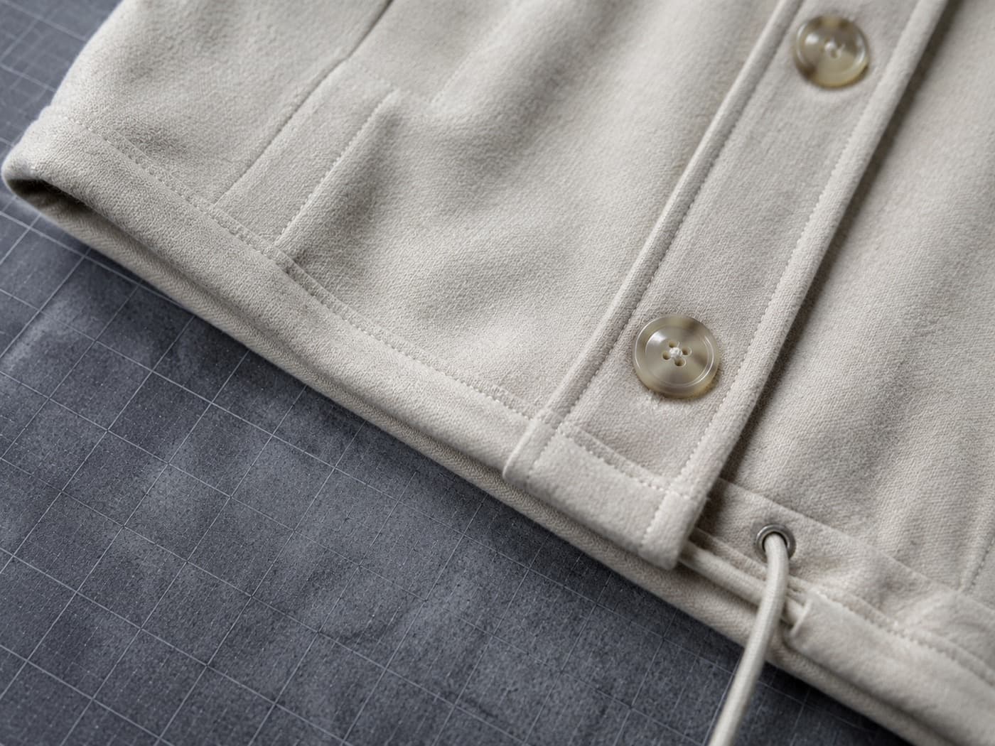 Close view of a hem edge, a button placket and a drawcord channel on pale wool, lit evenly on a dark plate.